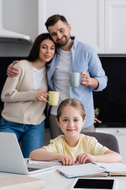 girl smiling at camera while doing homework near happy parents on blurred background - Photo, Image