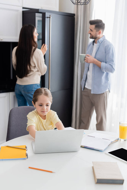 menina digitando no laptop ao fazer lição de casa perto dos pais falando sobre fundo borrado - Foto, Imagem
