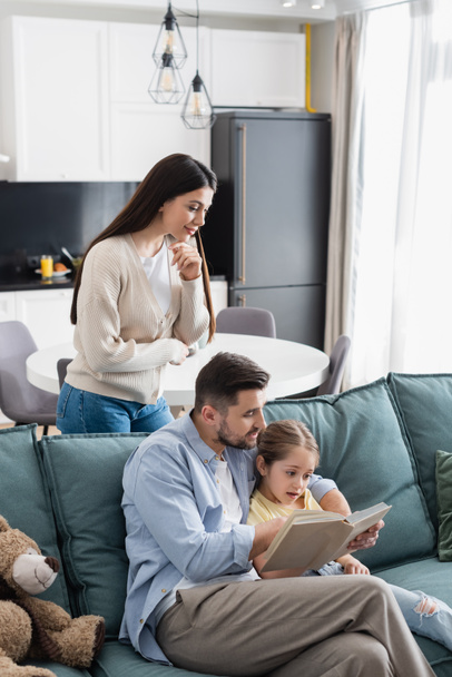 woman standing near husband and daughter reading book on sofa at home - Foto, immagini