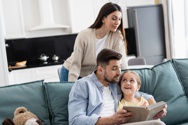 woman looking at happy daughter and husband reading book on couch - Φωτογραφία, εικόνα