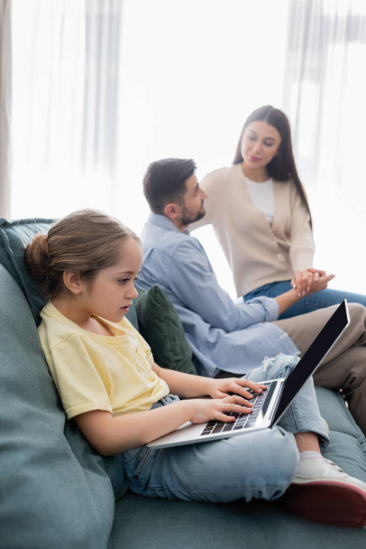 girl typing on laptop near parents talking and holding hands on blurred background - Photo, Image