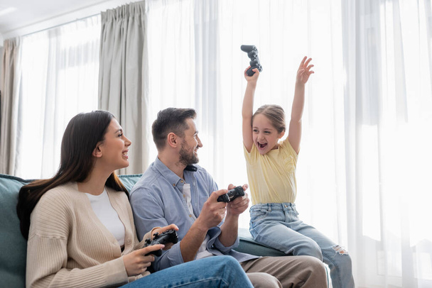 KYIV, UKRAINE - APRIL 8, 2021: cheerful girl with joystick showing win gesture near happy parents at home - Fotografie, Obrázek