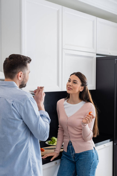 smiling woman looking at husband tasting meal in kitchen - Foto, Imagem
