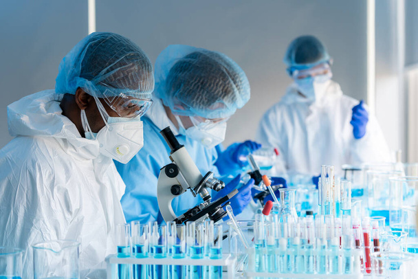 Οι Αφροαμερικανοί επιστήμονες χρησιμοποιούν μικροσκόπια για να ερευνήσουν το εμβόλιο του ιού covid-19 σε ιατρικό εργαστήριο. Ο γιατρός χρησιμοποιεί τη βιοτεχνολογία για την έρευνα του κορωνοϊού στο εργαστήριο χημείας. Φαρμακευτική επιστήμη. - Φωτογραφία, εικόνα