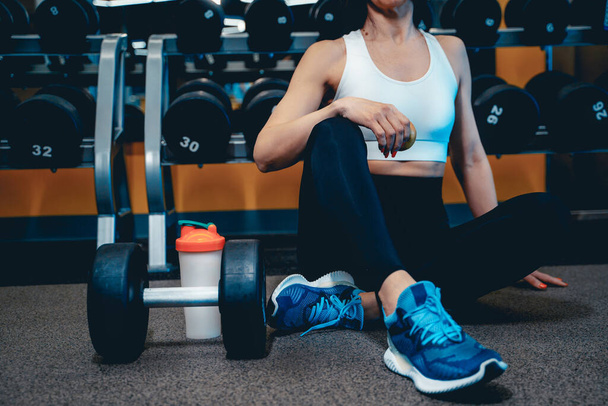 a girl in a sports uniform sits on the floor in the gym and rests, drinks a cocktail from a shaker on the background of a dumbbell - Foto, immagini