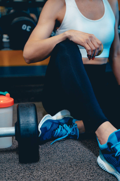 a girl in a sports uniform sits on the floor in the gym and rests, drinks a cocktail from a shaker - Foto, Imagem