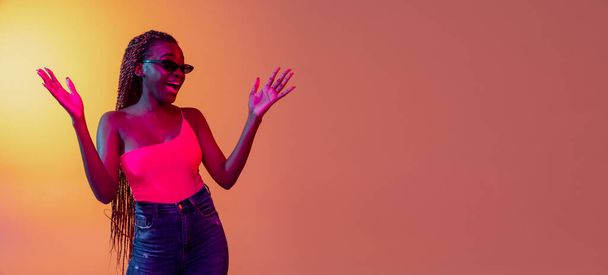 Young pretty African girl, female fashion model in sunglasses and white headphones isolated on yellow background in neon light. - Photo, Image