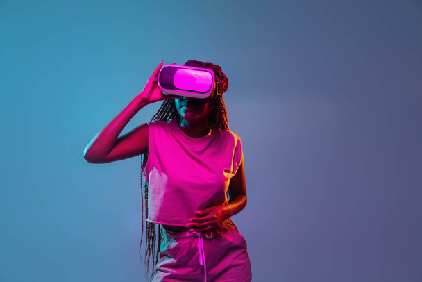 Portret van een jonge Afrikaanse vrouw met een vr bril op een blauwe neon achtergrond. Concept van virtual reality, entertainment en mensen. - Foto, afbeelding