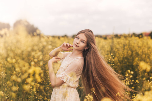 Young beautiful girl with long hair flying in the wind against the background of rapeseed field. Breeze playing with girl's hair - Fotoğraf, Görsel