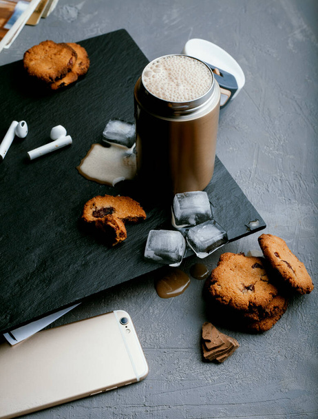 Холодный кофе в стальной термокружке, с миндальным молоком, кубиками льда и домашним печеньем на черном каменном фоне. Холодный летний напиток - Фото, изображение