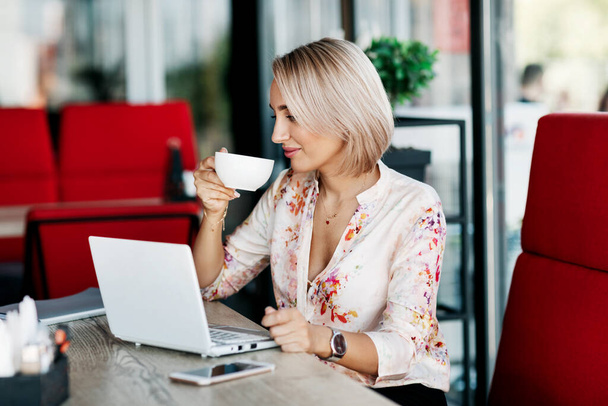 Una mujer de negocios elegante bebe café, trabaja en una cafetería, usa una computadora portátil, disfruta de un descanso. Negocios, negocios online, freelance - Foto, Imagen