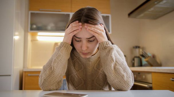 Perturbada e estressada adolescente sentada na cozinha e olhando para o smartphone - Foto, Imagem