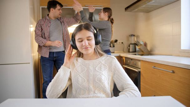 Teenage girl putting on headphones and enjoying music while parents shouting an arguing on the background. Family violence, conflicts and relationship problems - Photo, Image