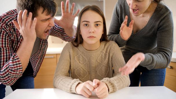 Father and mother shouting and screaming at teenage daughter sitting behind table on kitchen. Family violence, conflicts and relationship problems - Foto, Imagen