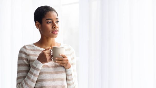 Young Black Lady Drinking Coffee While Standing Near Window At Home - 写真・画像