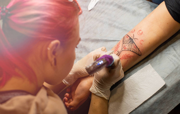 Tatuagem mestre desenha a tinta vermelha na tatuagem dos clientes. Tatuagem artista segurando uma máquina de tatuagem rosa em luvas estéreis pretas e trabalhando no tapete azul profissional. - Foto, Imagem