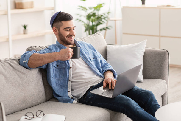 Sonriente hombre judío viendo vídeo en la computadora, bebiendo café caliente - Foto, imagen