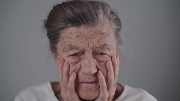 Mujer mayor de 90 años con arrugas profundas y cabello gris tiene estrés psicológico, llora y mira a la cámara sosteniendo su cara con las manos en el fondo gris. Depresión y soledad en ancianos - Metraje, vídeo