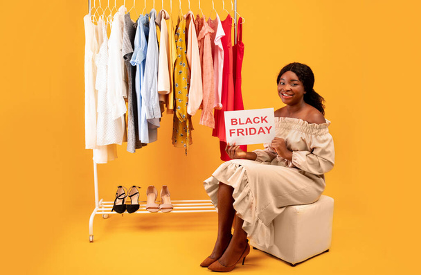 Czarna pani trzyma czarny znak FRIDAY, oferując ogromną sezonową sprzedaż, siedzi w pobliżu wieszak na ubrania na pomarańczowym tle - Zdjęcie, obraz