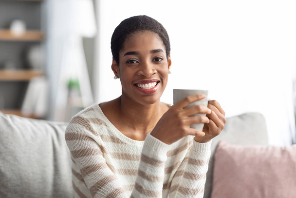 Home Freizeit. Lächelnde junge schwarze Frau sitzt auf Couch und trinkt Kaffee - Foto, Bild