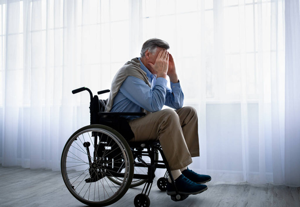 Пожилой инвалид в инвалидной коляске закрывает лицо руками, плачет у окон, страдает от депрессии в помещении - Фото, изображение