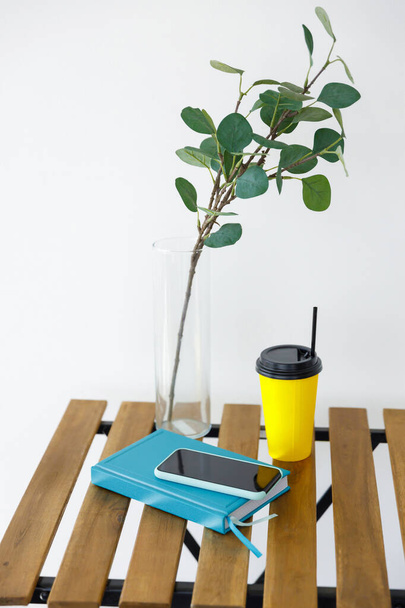 Wooden table against background of white wall. Vase with eucalyptus branch, yellow paper coffee cup and smartphone with notepad. Freelancer workplace - Valokuva, kuva