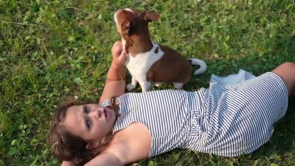 милая маленькая кавказская девочка ласкает собаку, лежащую на траве - Кадры, видео