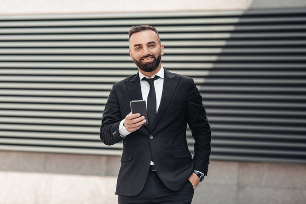 ポケットに手を入れてオフィスビルの近くに立つスマートフォンを使って黒いスーツを着た幸せな成熟した起業家 - 写真・画像