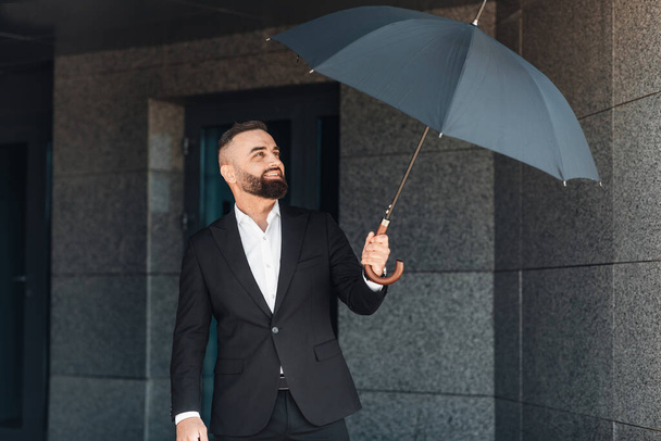 Mature office worker wearing classical suit holding umbrella while walking outdoors, going home after work in office - Foto, Imagen