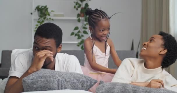 Afro american family little funny daughter child girl kid tickles playing with mom lying on couch having fun at home black african father suffering from allergies pain in eyes poor vision rubs eye - Footage, Video
