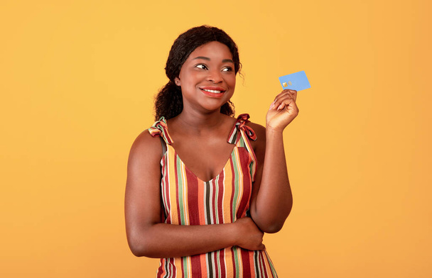Koncept bezhotovostní platby. Veselý černošky žena drží kreditní kartu v ruce přes oranžové studio pozadí - Fotografie, Obrázek
