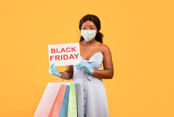 Black woman wearing face mask, holding BLACK FRIDAY sign, holding shopping bags, purchasing things during coronavirus - Photo, Image
