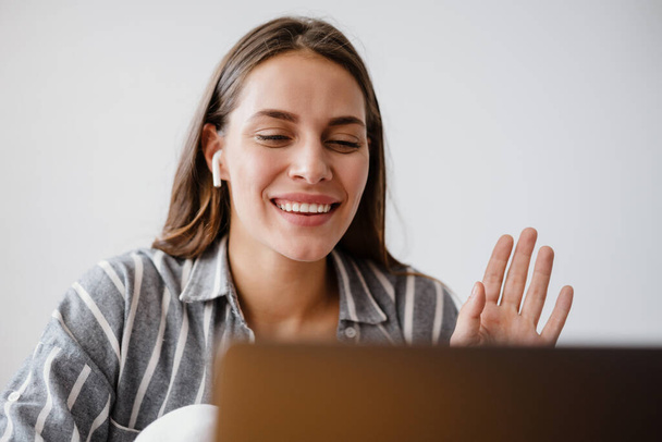 Cheerful beautiful woman in earphones waving hand while using laptop at home - Fotó, kép