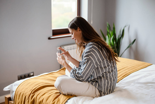 Pleased brunette woman using cellphone while sitting on bed at home - Foto, imagen