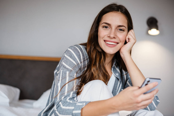 Pleased brunette woman smiling and using cellphone while sitting on bed at home - Foto, Imagem