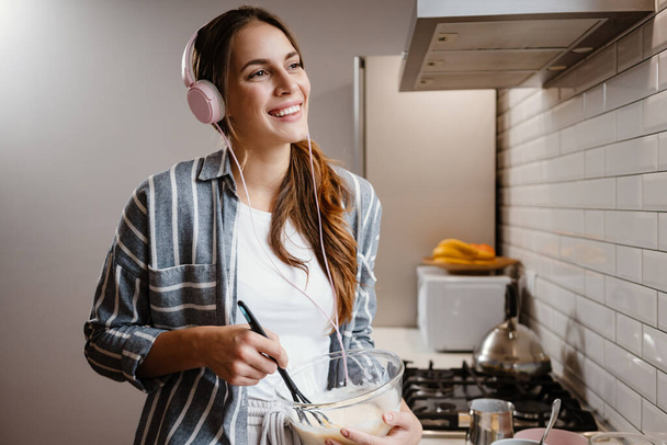 Задоволена красива жінка в навушниках посміхається і робить млинці тістом на домашній кухні
 - Фото, зображення