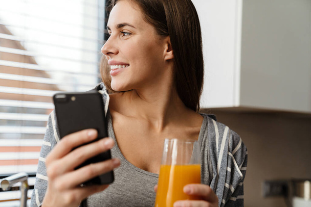 Okouzlující veselá žena pití šťávy při použití mobilního telefonu v útulné kuchyni doma - Fotografie, Obrázek