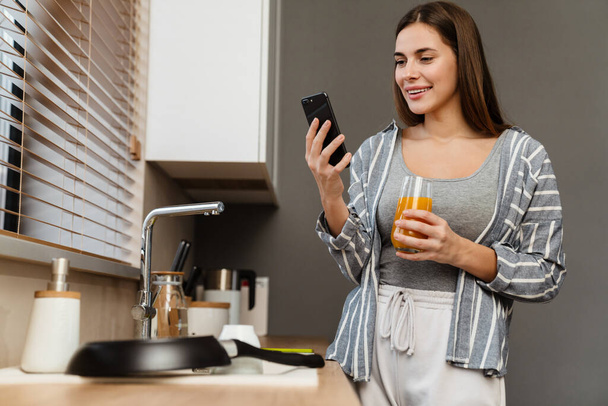 Чарівна весела жінка п'є сік, використовуючи мобільний телефон у затишній кухні вдома
 - Фото, зображення