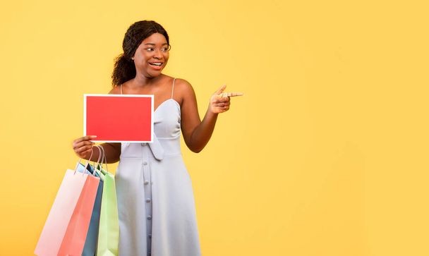 Junge schwarze Frau hält Einkaufstüten und leeres Schild mit Attrappe in der Hand und zeigt auf Leerstelle vor orangefarbenem Hintergrund - Foto, Bild