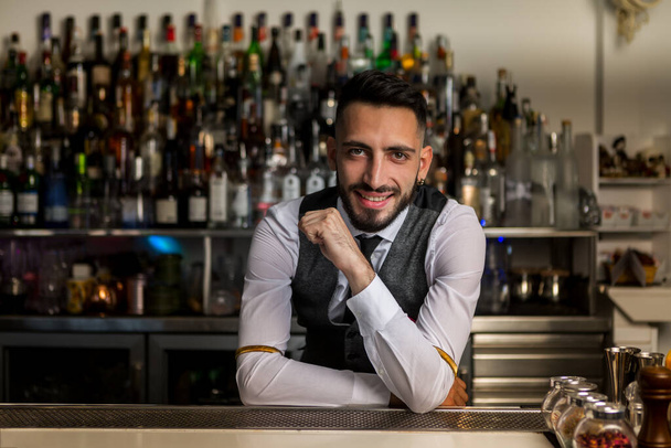 Handsome young bearded Hispanic male barkeeper in elegant clothes smiling friendly and looking at camera while standing behind bar counter with alcoholic drinks - Foto, immagini