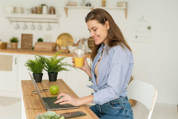 幸せな美しい若い女性が自宅でテーブルに座って、ノートパソコンで作業し、おいしいオレンジジュースを飲んでいます。検疫条件下における家庭からの仕事と研究 - 写真・画像