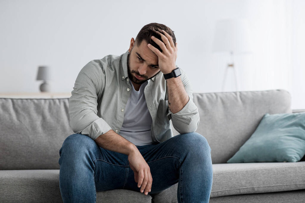 Nešťastný muž se dotýká vlasů, sedí doma na pohovce a přemýšlí o problémech - Fotografie, Obrázek