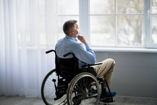Handicappato vecchio sulla sedia a rotelle indossando maschera facciale, che soffre di solitudine in casa di riposo durante covid - Foto, immagini