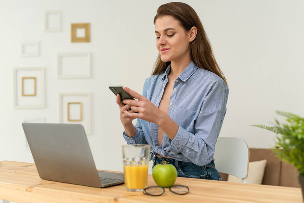 Inteligentna kobieta siedzi przy stole kuchennym, szukając plików w telefonie i wysyłając je do laptopa przez domową sieć bezprzewodową. Młoda piękna kobieta freelancer synchronizuje - Zdjęcie, obraz