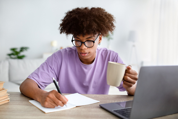Εστιασμένη μαύρη έφηβη με φλιτζάνι καφέ κρατώντας σημειώσεις κατά τη διάρκεια της online διάλεξης για το φορητό υπολογιστή στο σπίτι - Φωτογραφία, εικόνα
