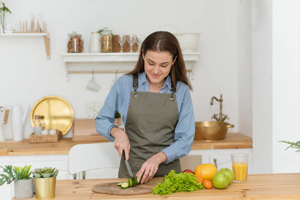 Zielona dieta, kobieta krojąca ogórki. Piękna kobieta przygotowuje smaczną sałatkę warzywną w kuchni w domu - Zdjęcie, obraz
