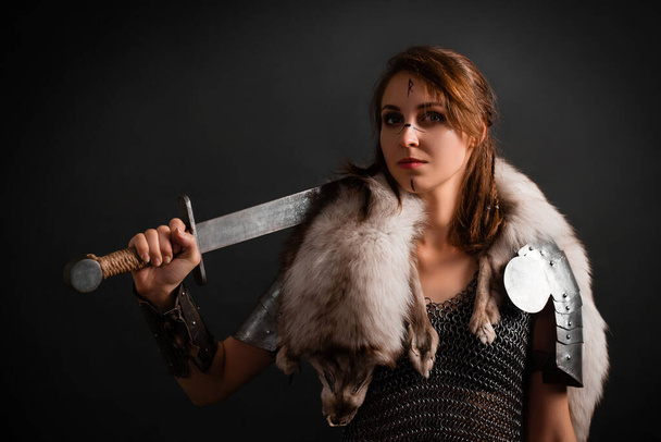 Portrait of a medieval woman warrior in chain mail armor and polar fox fur on her shoulders standing with a sword in hand on a dark background.  Female warrior silhouette. - Photo, image
