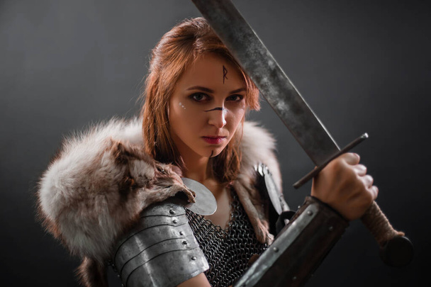 Portrait of a medieval woman warrior in chain mail armor and polar fox fur on her shoulders standing with a sword in hand on a dark background.  Female warrior silhouette. - Foto, Imagem