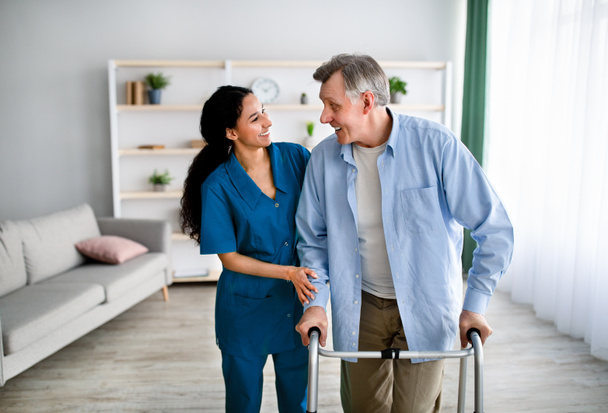 Vrouwelijke verzorger helpt oudere man om thuis met frame te lopen. Professionele gezondheidszorg voor senioren - Foto, afbeelding