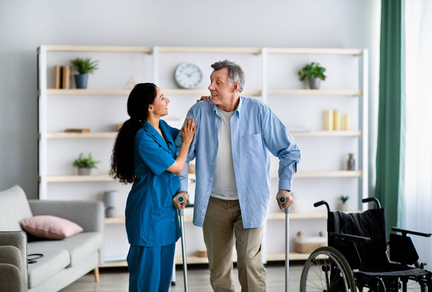 Female nurse helping elderly male to walk with crutches at home. Professional medical care for disabled older patients - Photo, Image